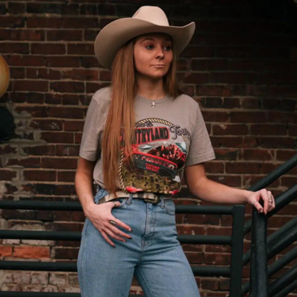 Woman wearing a cowboy hat, graphic t-shirt, and jeans leaning against a railing.
