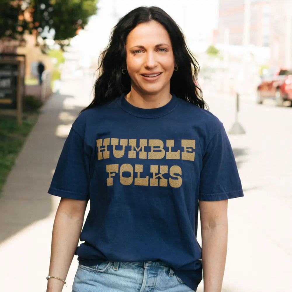 Woman wearing a navy blue t-shirt with ’HUMBLE FOLKS’ printed in gold letters.