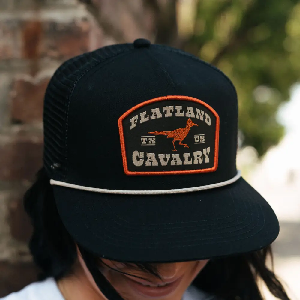 Black baseball cap with an orange and white ’Flatland Cavalry’ patch featuring a bird silhouette.